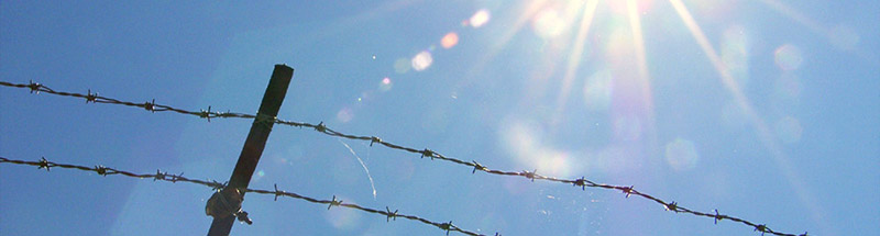 Blauer Himmel mit Sonnenstrahlen über einem Staheldrahtzaun